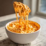 A spoonful of hot, gooey chili cheese dip is lifted from a white bowl, showing long, cheesy strings.