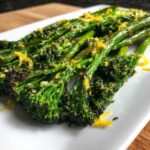 Close-up of bright green, roasted broccolini topped with lemon zest and seasoning on a white plate.