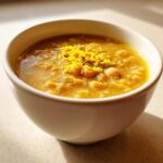 Close-up of a hearty, yellow chickpea soup garnished with bright yellow lemon zest in a white bowl.