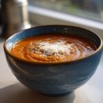 Close-up of a bowl of rich tomato basil soup topped with shredded Parmesan and herbs.