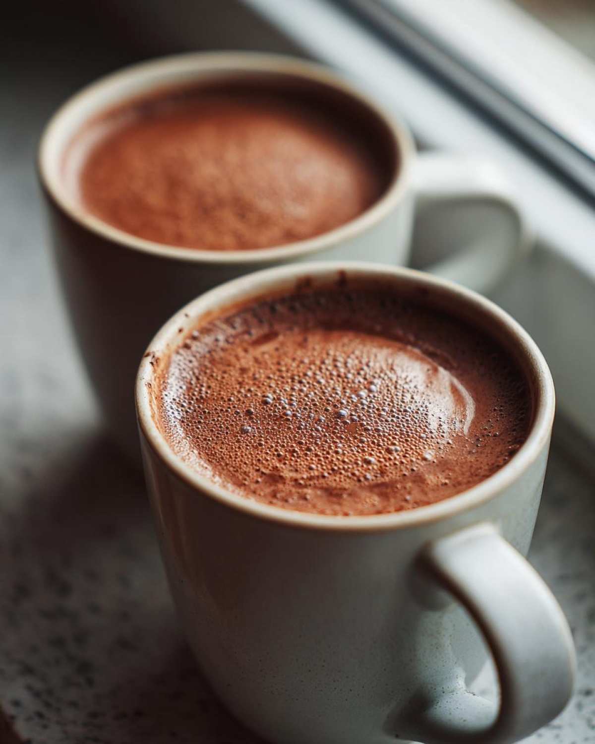 Close-up of two creamy mugs filled with rich hot chocolate, sitting by a window.