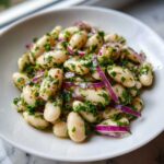 A close-up, appetizing shot of a vibrant white bean salad mixed with thinly sliced red onion and fresh parsley.