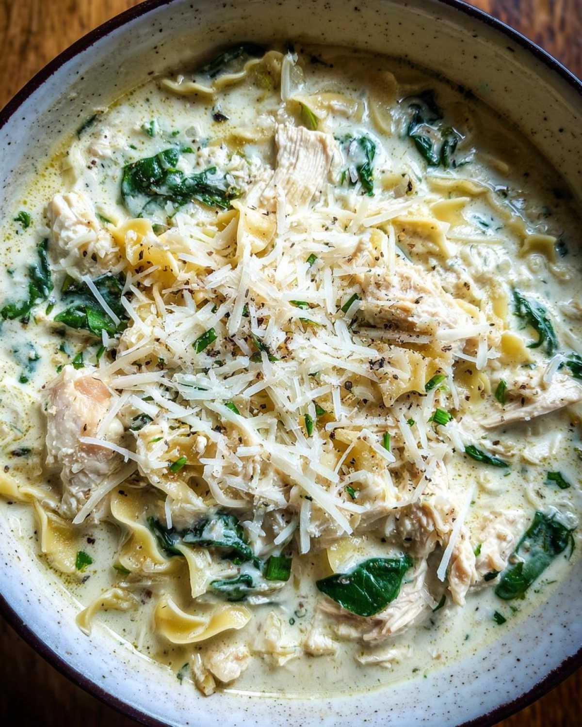 A close-up, top-down view of a bowl of creamy white chicken lasagna soup topped with shredded Parmesan cheese and black pepper.