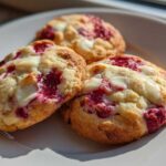 Three freshly baked white chocolate raspberry cookies resting on a white plate, bathed in sunlight.