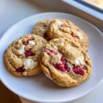 Close-up of four freshly baked white chocolate raspberry cookies resting on a white plate.