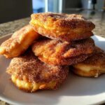A stack of freshly made, golden brown buñuelos heavily coated in cinnamon sugar, resting on a white plate.
