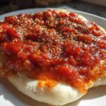 Close-up of thick, rich pizza sauce topped with dried herbs spread over a piece of uncooked or lightly baked dough.