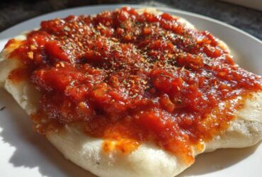 Close-up of thick, rich pizza sauce topped with dried herbs spread over a piece of uncooked or lightly baked dough.