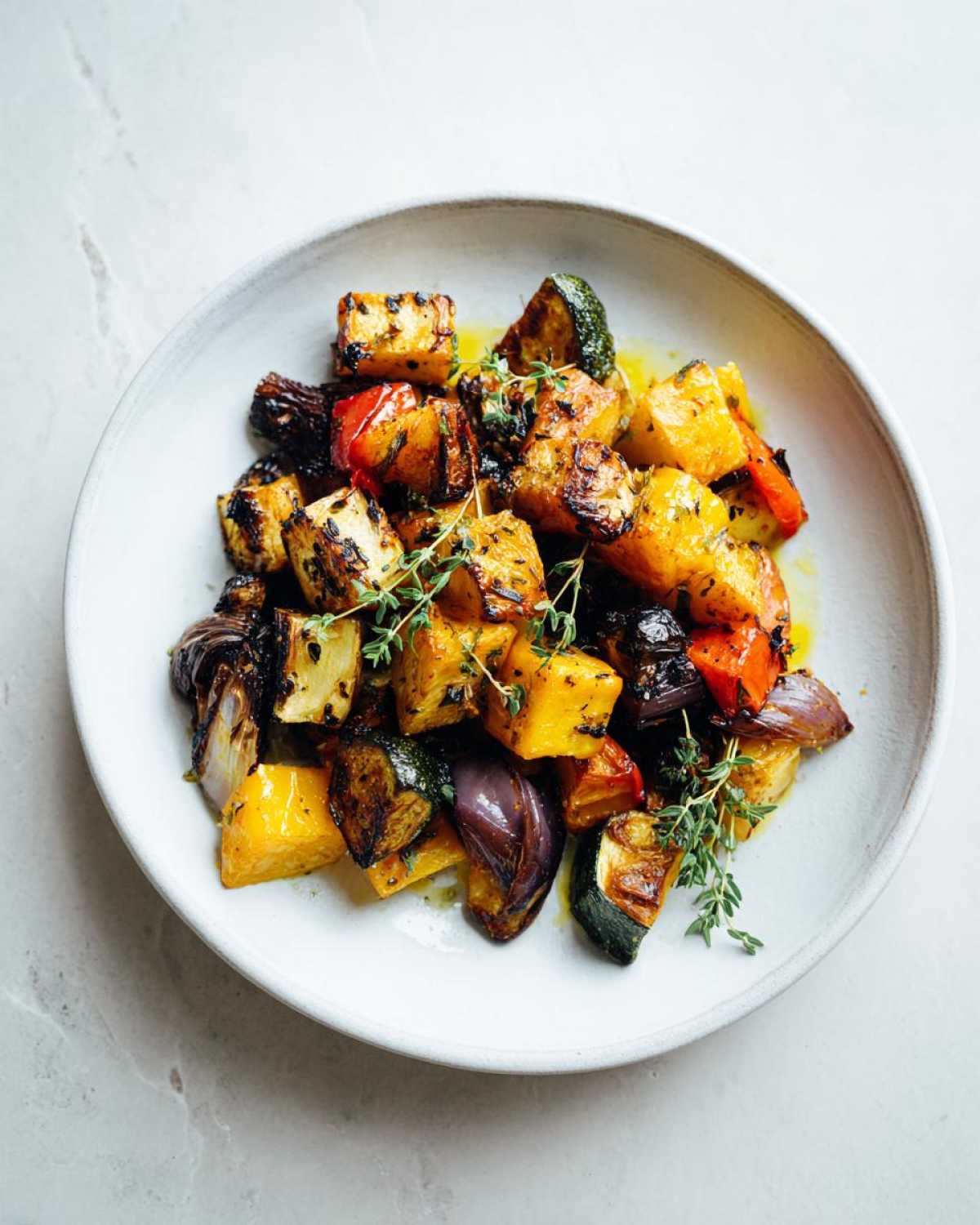 A bowl of colorful, caramelized roasted vegetables, including squash, zucchini, and red onion, drizzled with oil and topped with fresh thyme.