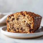 A close-up of a freshly baked vegan banana bread loaf studded with chocolate chips, sitting on a small plate.