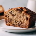 Close-up of a thick slice of moist vegan banana bread loaded with chocolate chips, served on a white plate.