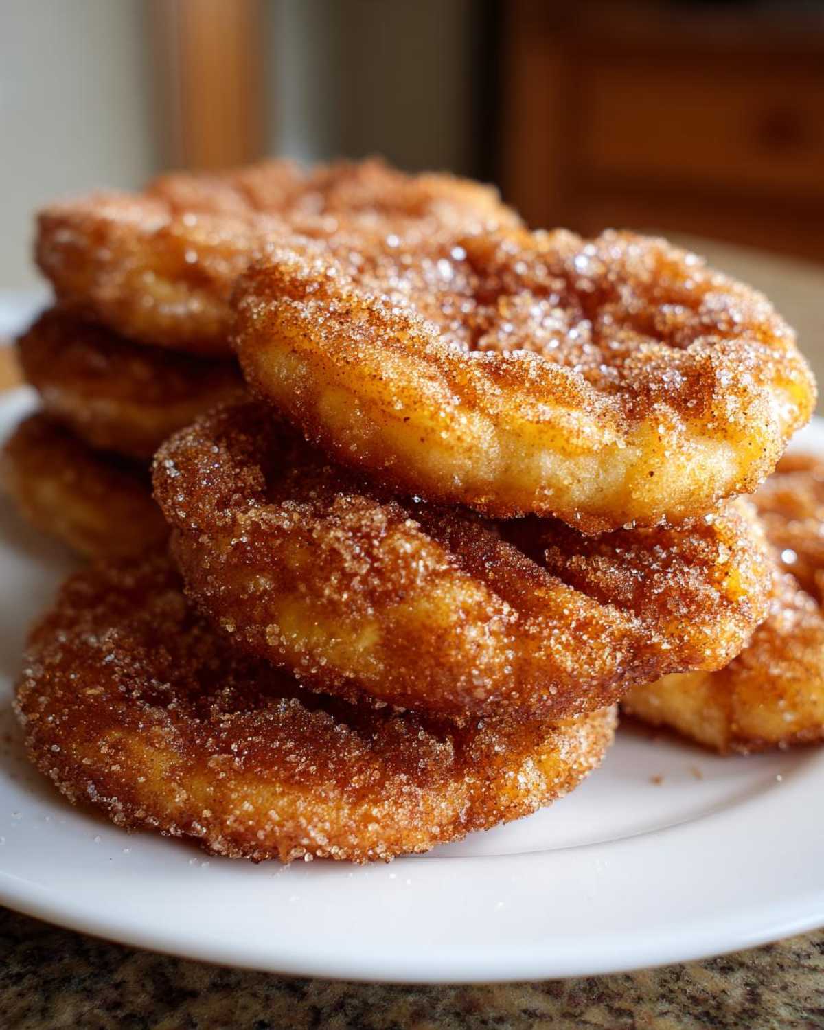 A close-up stack of golden-brown buñuelos generously coated in sparkling cinnamon sugar, resting on a white plate.