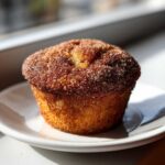 A single, golden-brown cinnamon sugar french toast muffin coated in sparkling sugar, sitting on a white plate.