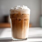 A tall glass filled with a Copycat Starbucks Iced White Chocolate Mocha, layered with coffee, milk, and topped with whipped cream.