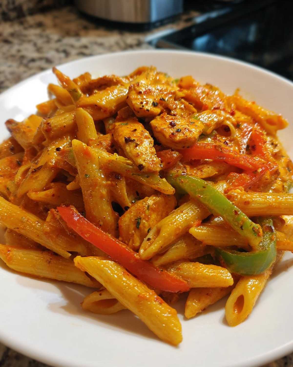 A close-up of a white bowl filled with creamy, orange-hued Rasta Pasta mixed with seasoned chicken pieces and sliced red and green bell peppers.