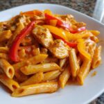 A close-up shot of creamy Rasta Pasta featuring penne, seasoned chicken pieces, and bright red and yellow bell pepper strips.