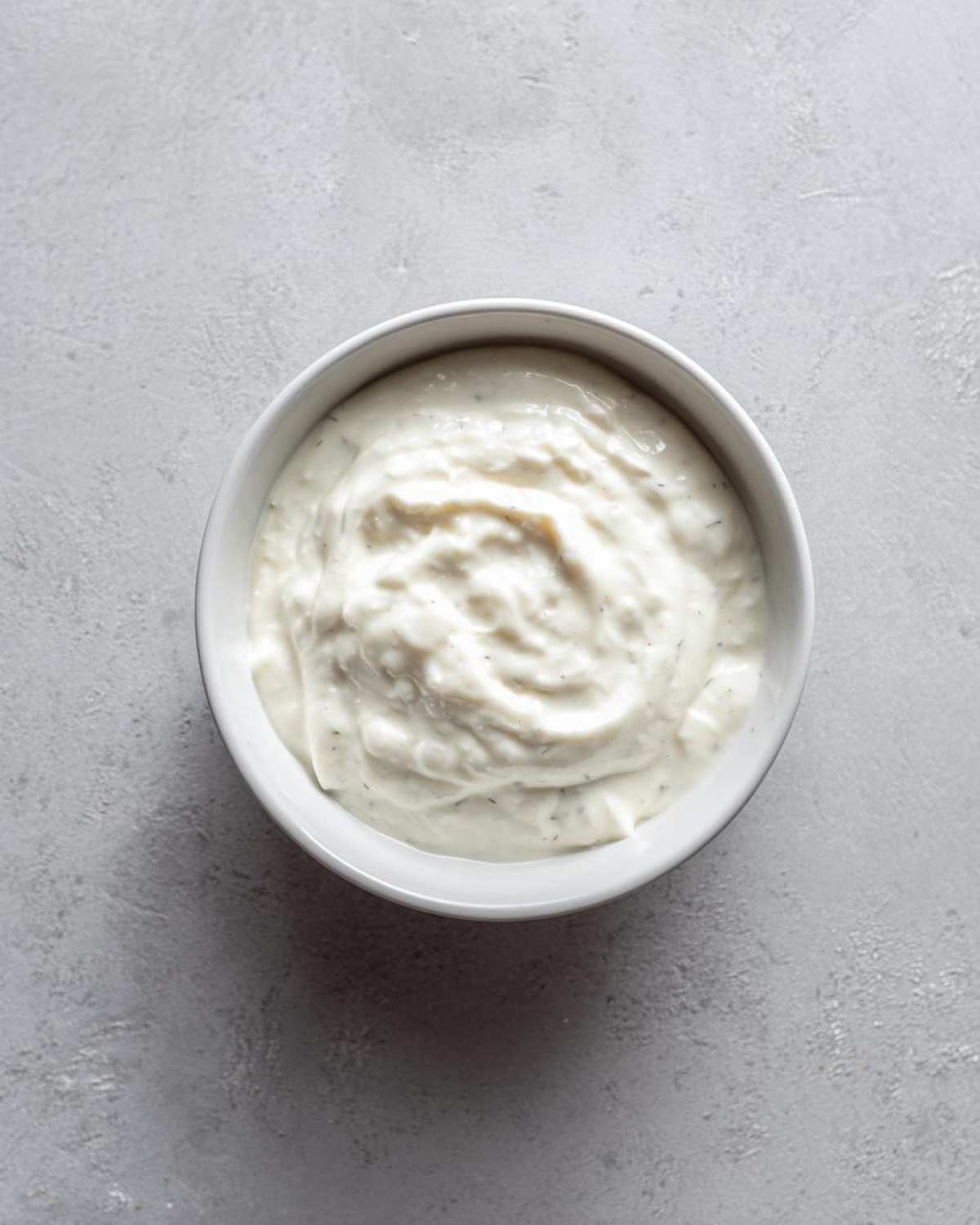 Overhead view of a white bowl filled with thick, creamy horseradish sauce speckled with green herbs.