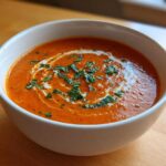 A close-up of a white bowl filled with rich, orange tomato bisque, swirled with cream and topped with fresh basil.