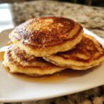 A stack of three fluffy coconut flour pancakes drizzled generously with golden syrup on a white plate.