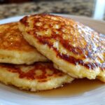 A stack of three fluffy coconut flour pancakes drizzled generously with golden syrup on a white plate.