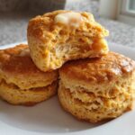 Three fluffy sweet potato biscuits stacked on a white plate, one broken open with melting butter on top.