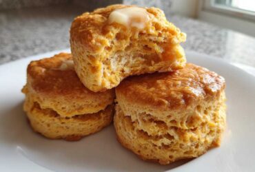Three fluffy sweet potato biscuits stacked on a white plate, one broken open with melting butter on top.