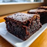 A close-up, fudgy square of dark brown gingerbread brownies resting on a white rectangular plate.