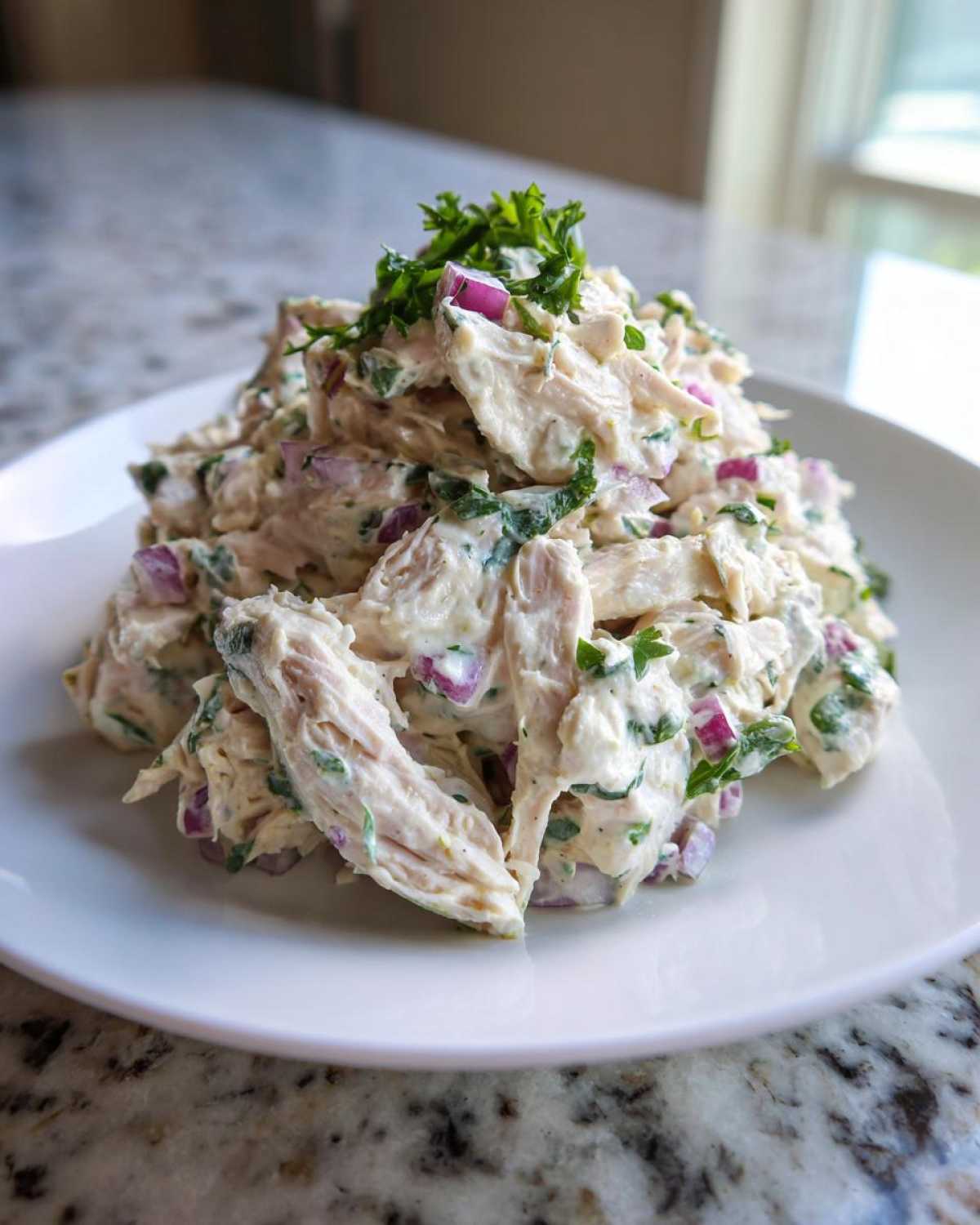 A mound of creamy greek yogurt chicken salad mixed with red onion and parsley, served on a white plate.