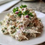 A mound of creamy greek yogurt chicken salad mixed with red onion and fresh parsley on a white plate.