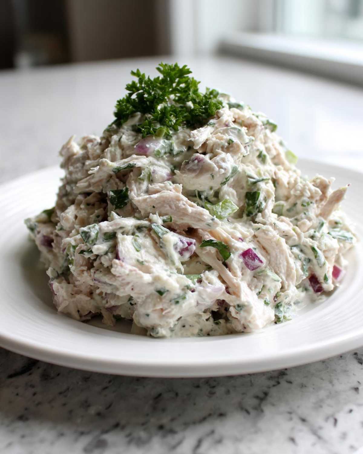 A mound of creamy greek yogurt chicken salad mixed with red onion and herbs, topped with fresh parsley.
