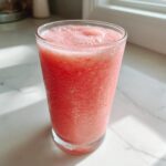 A tall glass filled with a bright pink, frothy watermelon smoothie sitting on a white marble counter.