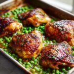Close-up of glossy, browned Sheet Pan Lemon Garlic Butter Chicken Legs resting on a bed of bright green peas.
