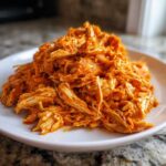 Close-up of shredded buffalo chicken piled high on a white plate, coated in orange sauce.