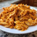 A close-up of moist, shredded buffalo chicken coated in bright orange sauce piled on a white plate.