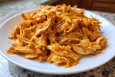 A close-up of moist, shredded buffalo chicken coated in bright orange sauce piled on a white plate.