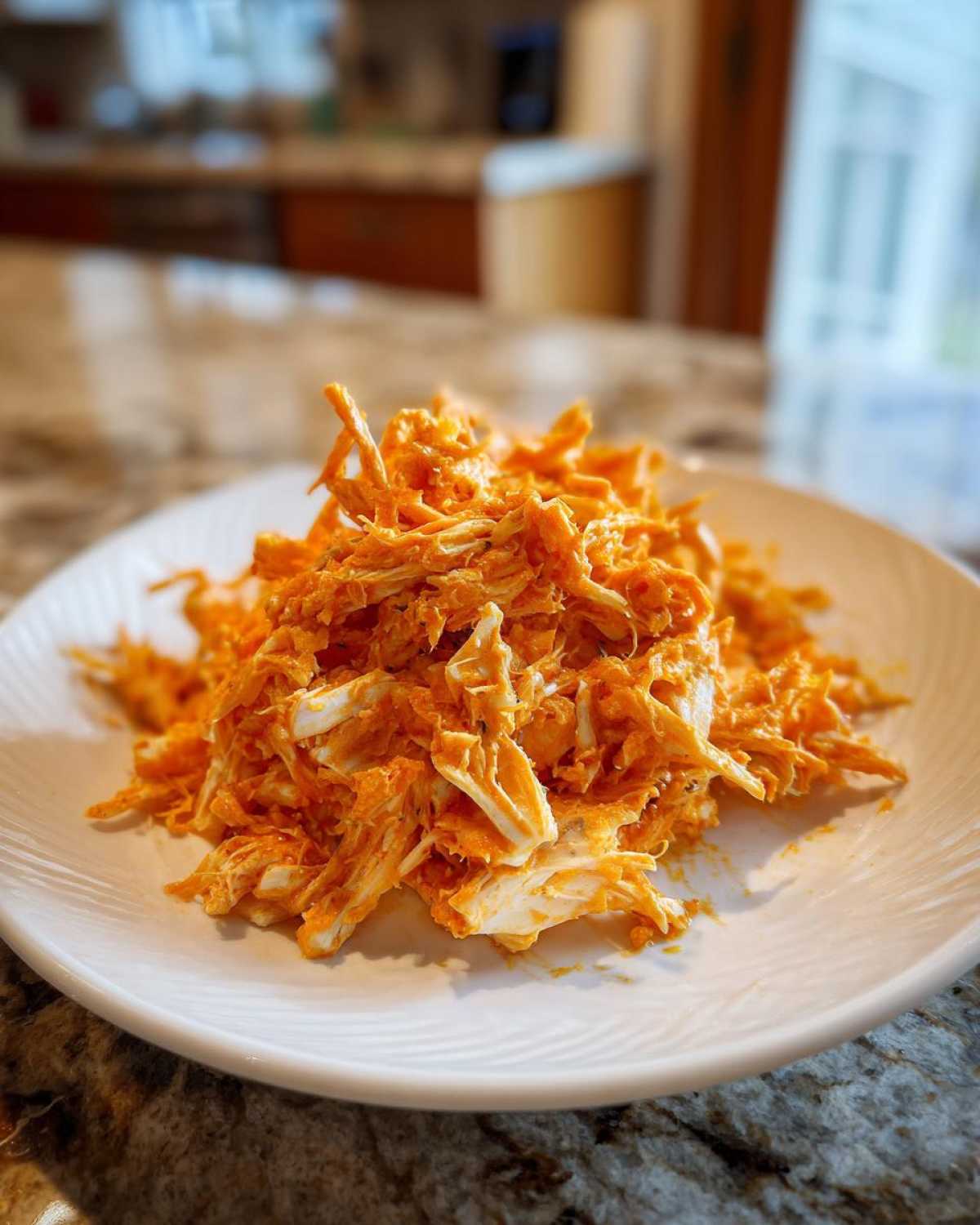 A close-up of shredded buffalo chicken coated in bright orange sauce piled on a white plate.