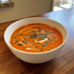 A close-up of a white bowl filled with rich, orange-red tomato bisque, swirled with cream and topped with fresh basil.