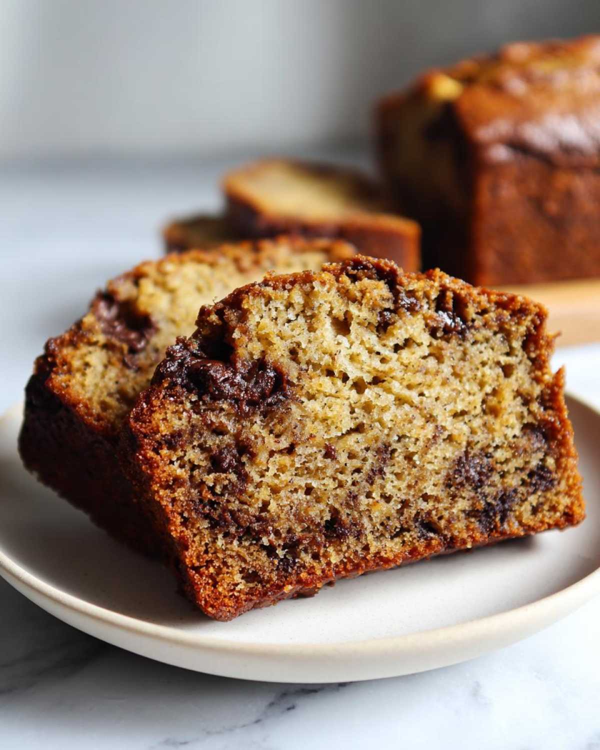 Two thick slices of moist vegan banana bread, rich with melted chocolate chunks, resting on a small plate.