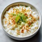 A bowl of Vegan Creamy Coconut Rice Pudding topped with toasted coconut flakes and lime zest/slices.