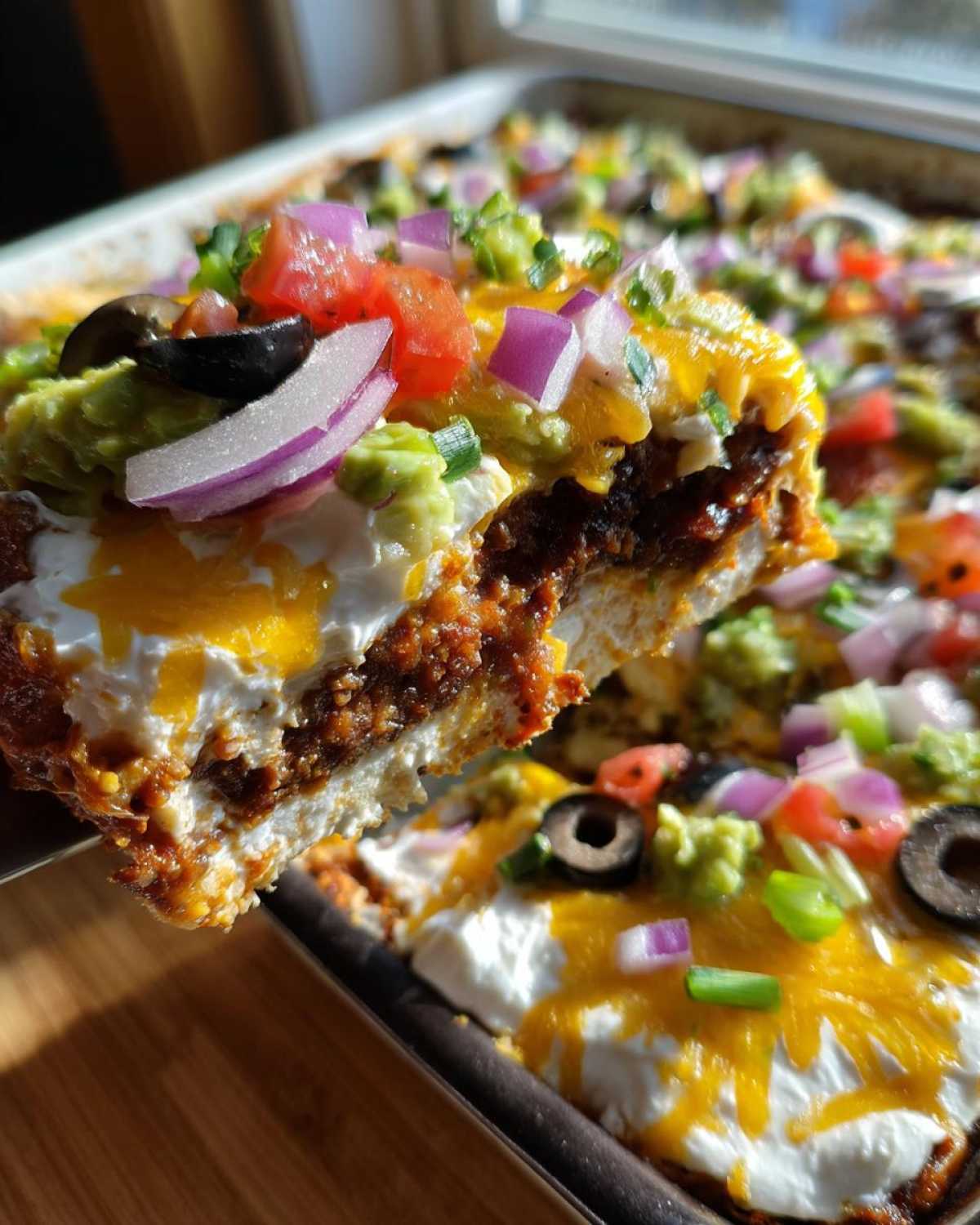 A spatula lifts a thick, layered slice of Avocado Party Dip topped with sour cream, cheese, onions, and olives.