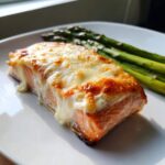 A perfectly cooked fillet of Baked Boursin Salmon topped with melted, slightly browned cheese, served alongside green asparagus.