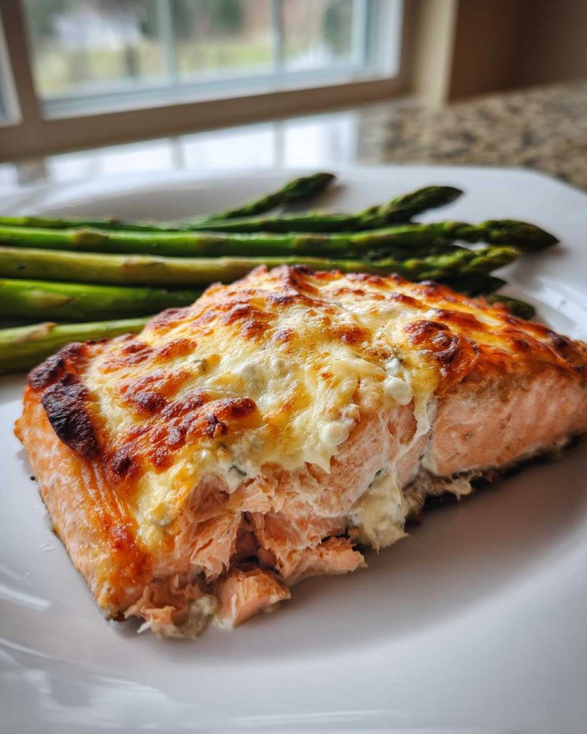 A close-up of flaky Baked Boursin Salmon topped with melted, browned cheese, served alongside roasted asparagus.