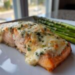 A perfectly cooked fillet of Baked Boursin Salmon topped with melted, creamy cheese and fresh parsley, served alongside bright green asparagus.