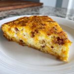 A close-up slice of Baked Hash Brown Quiche featuring a golden, cheesy top and visible bacon pieces.