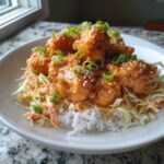 A close-up of a Bang Bang Salmon Bites Bowl featuring crispy salmon bites coated in orange sauce over rice and slaw, topped with sesame seeds and green onions.