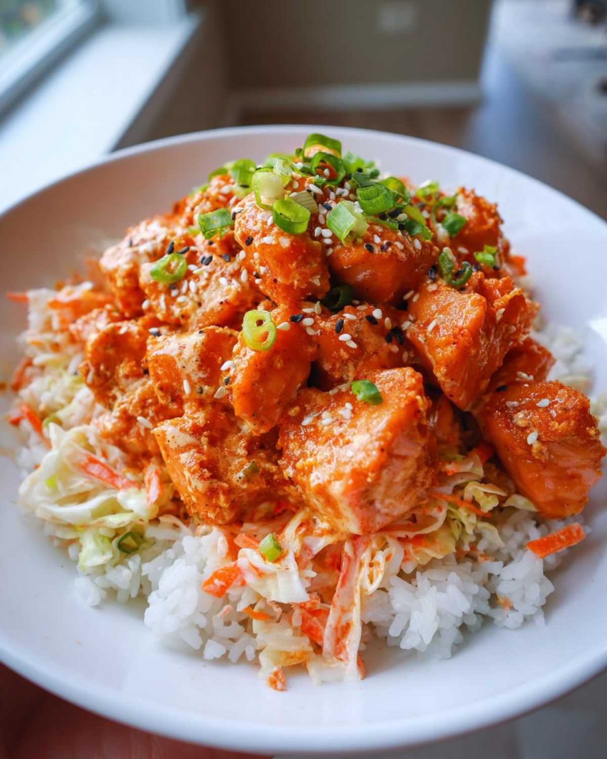 A close-up of a white bowl filled with Bang Bang Salmon Bites Bowl, featuring saucy salmon over rice and slaw, topped with green onions and sesame seeds.