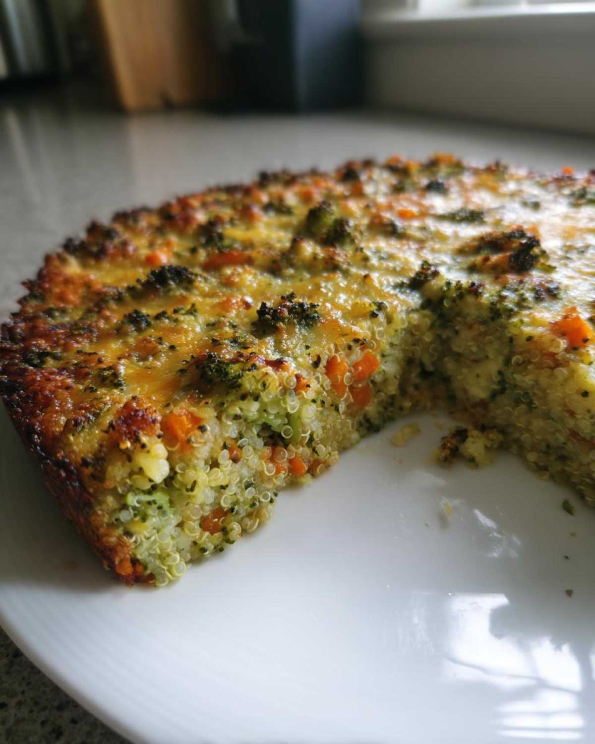 A close-up of a slice removed from a baked Cheesy Veggie Quinoa Casserole, showing quinoa, broccoli, and carrots inside with a golden cheese topping.