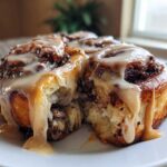 A close-up of two soft Cookie Butter Cinnamon Rolls dripping with thick, light brown icing on a white plate.