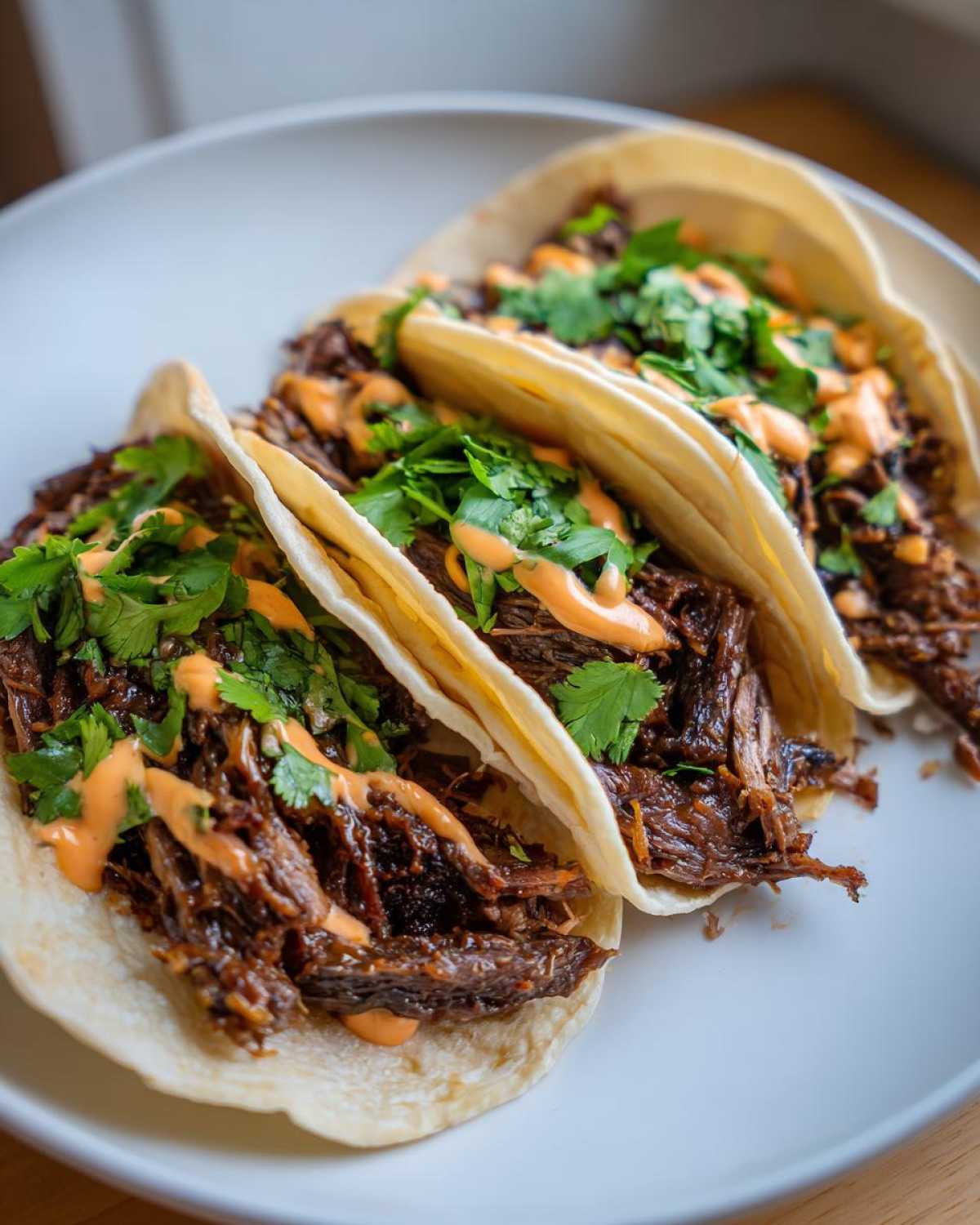 Three soft corn tortillas filled with Crockpot Korean Beef Tacos, topped with orange sauce and fresh cilantro.