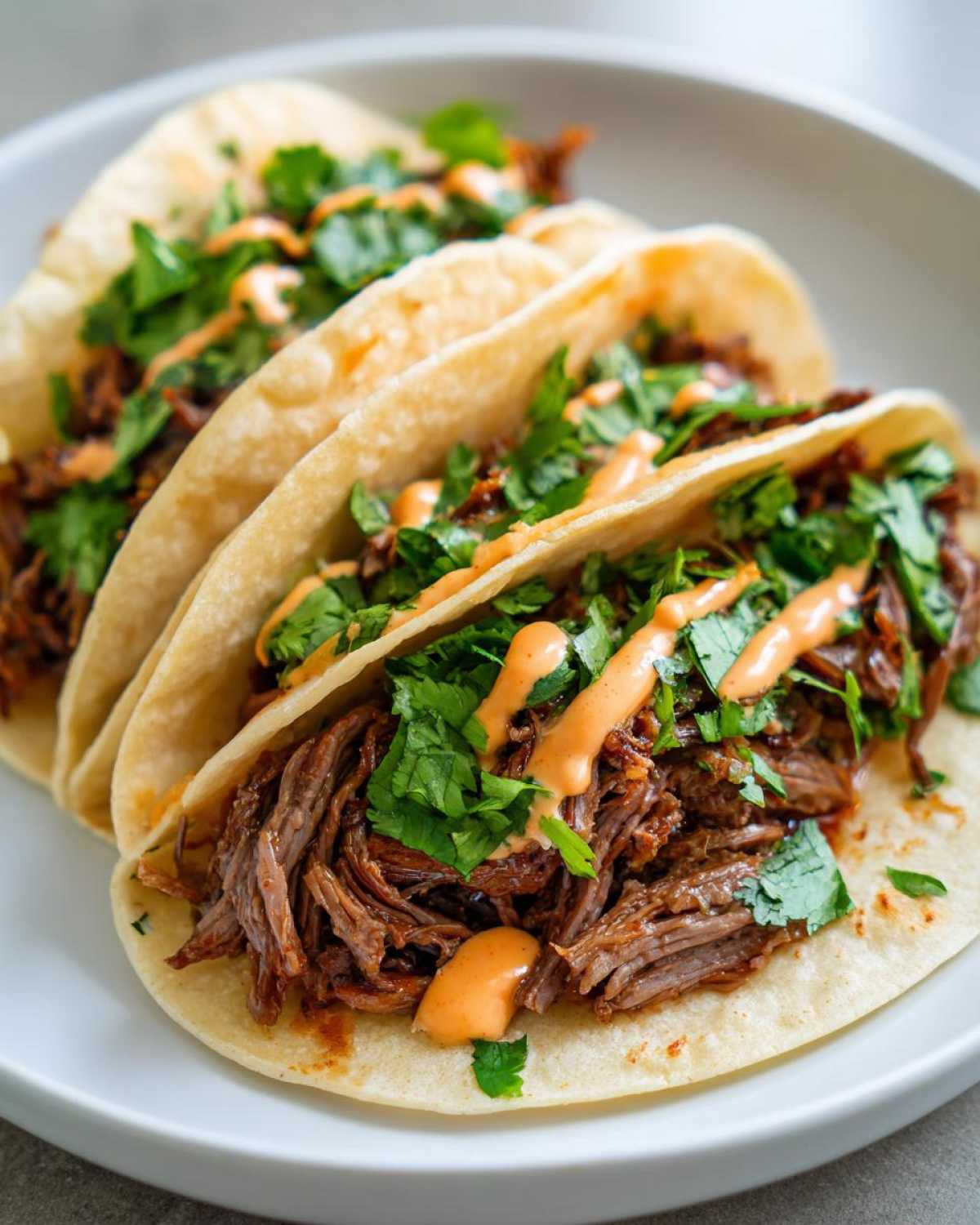 Three soft shell tacos filled with shredded Crockpot Korean Beef Tacos, topped with fresh cilantro and a drizzle of spicy orange sauce.